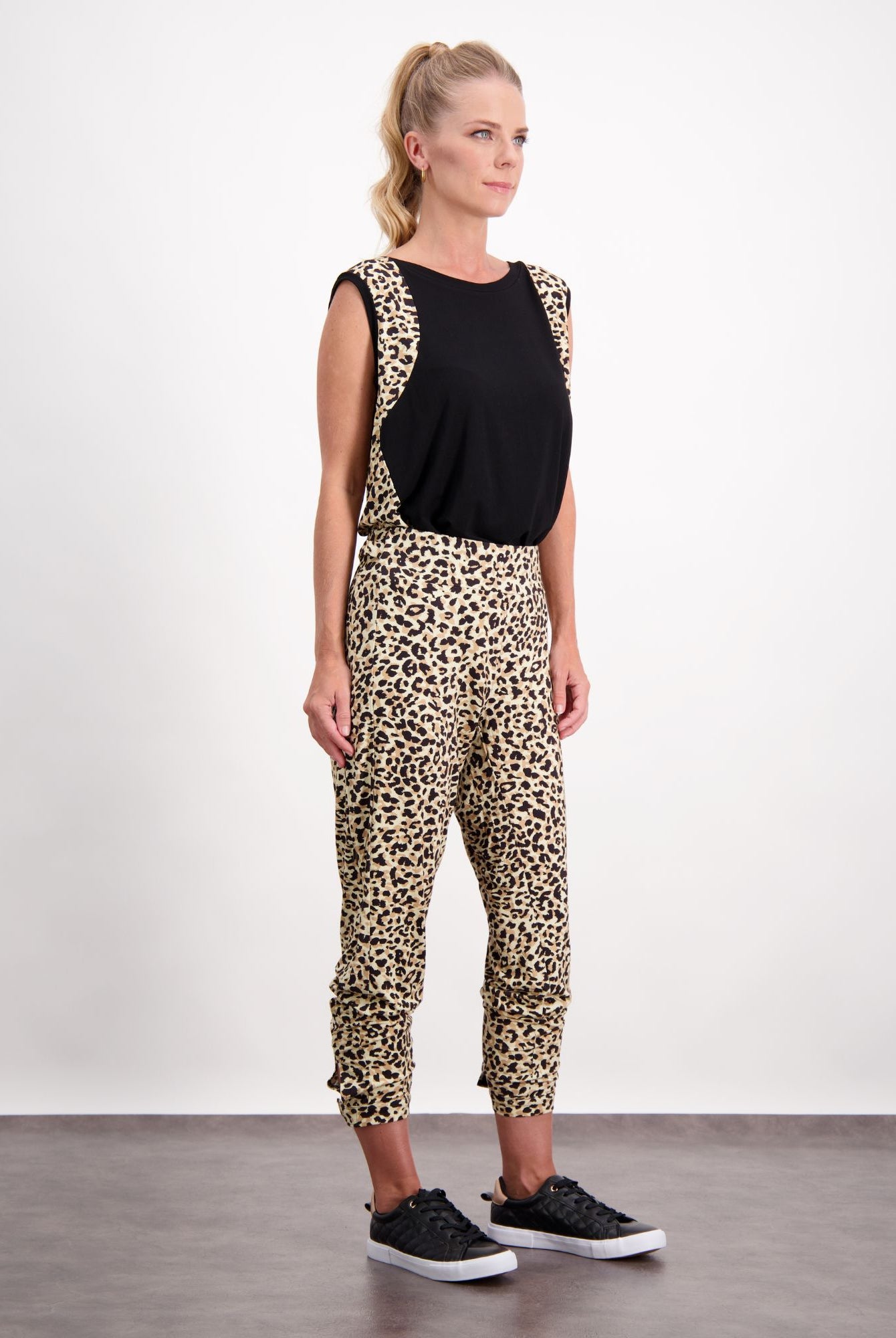 Lauren Sleeveless Physio Top shown in black, paired with leopard print pants, creating a stylish athletic ensemble. The sleeveless design features a classic round neckline and relaxed fit, perfect for exercise or casual wear. Model stands against white background wearing black sneakers.
