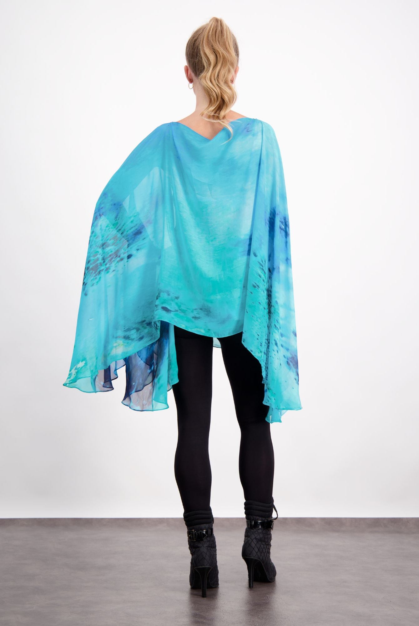 Oceania Kaftan shown from behind, featuring flowing aqua blue silk with darker blue watercolor patterns, draped elegantly over black leggings. The luxurious fabric creates a dramatic, wing-like silhouette against a white background.