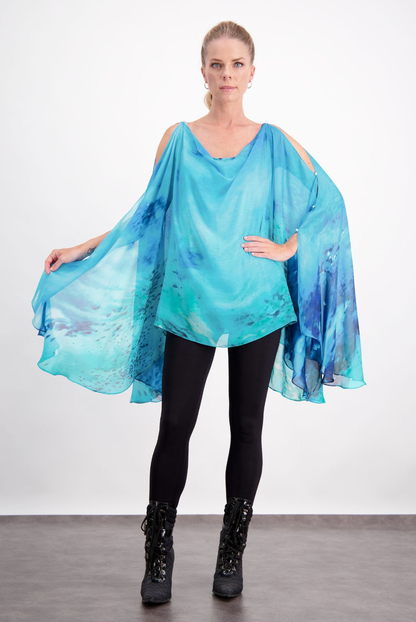 Oceania Kaftan featuring flowing silk fabric in vibrant aqua and deep blue watercolor design, with elegant cold-shoulder cut and dramatic cape-like draping, paired with black leggings and boots against a white background