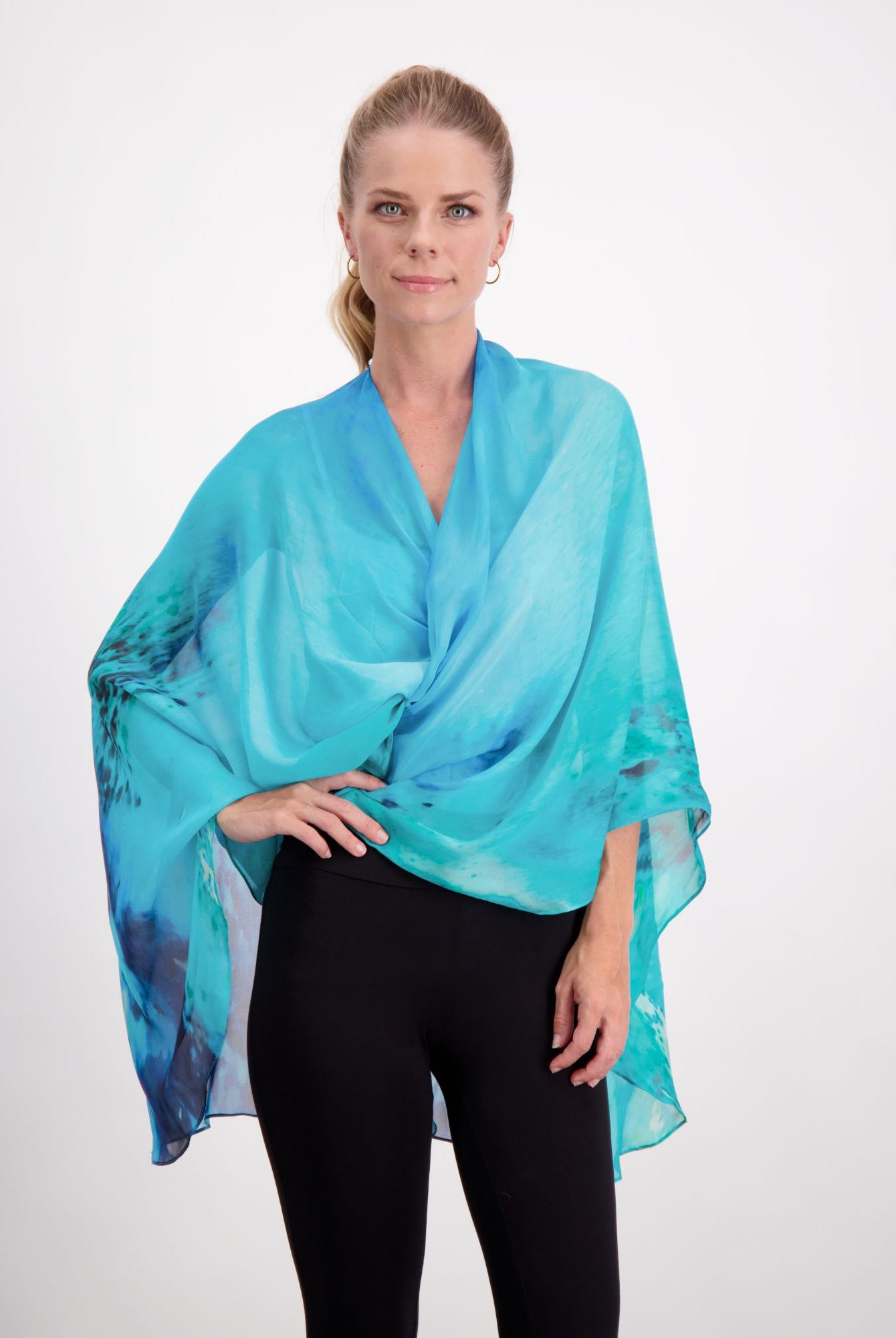 Oceania Kaftan displayed as an elegant aqua blue silk wrap with navy abstract patterns draped stylishly over black pants, showcasing its versatile styling potential and luxurious flowing fabric against a white background