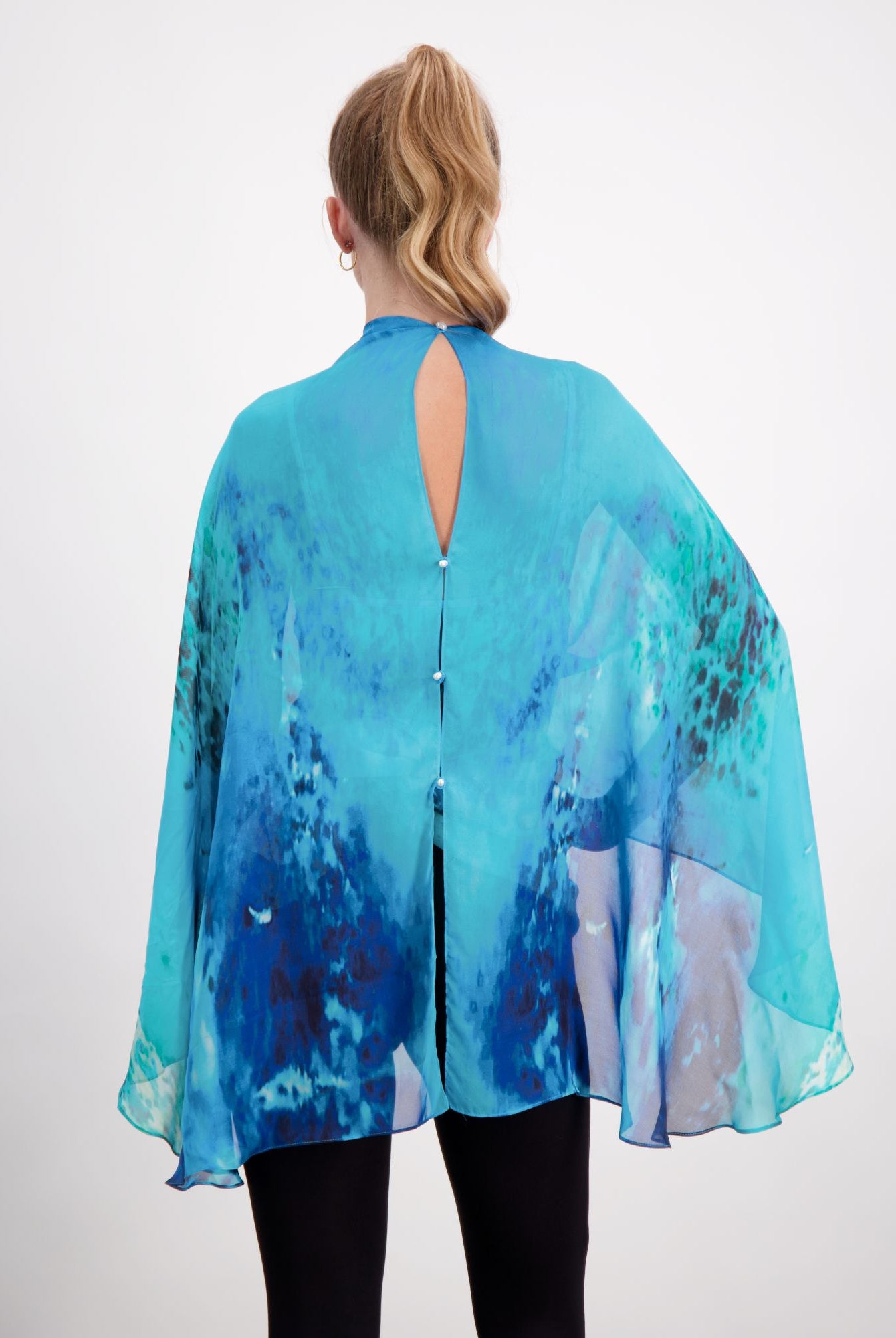 Back view of Oceania Kaftan featuring flowing silk fabric in vibrant aqua and deep blue watercolor patterns, with elegant keyhole button closure and dramatic cape-like silhouette against white background. Black pants complete the ensemble.