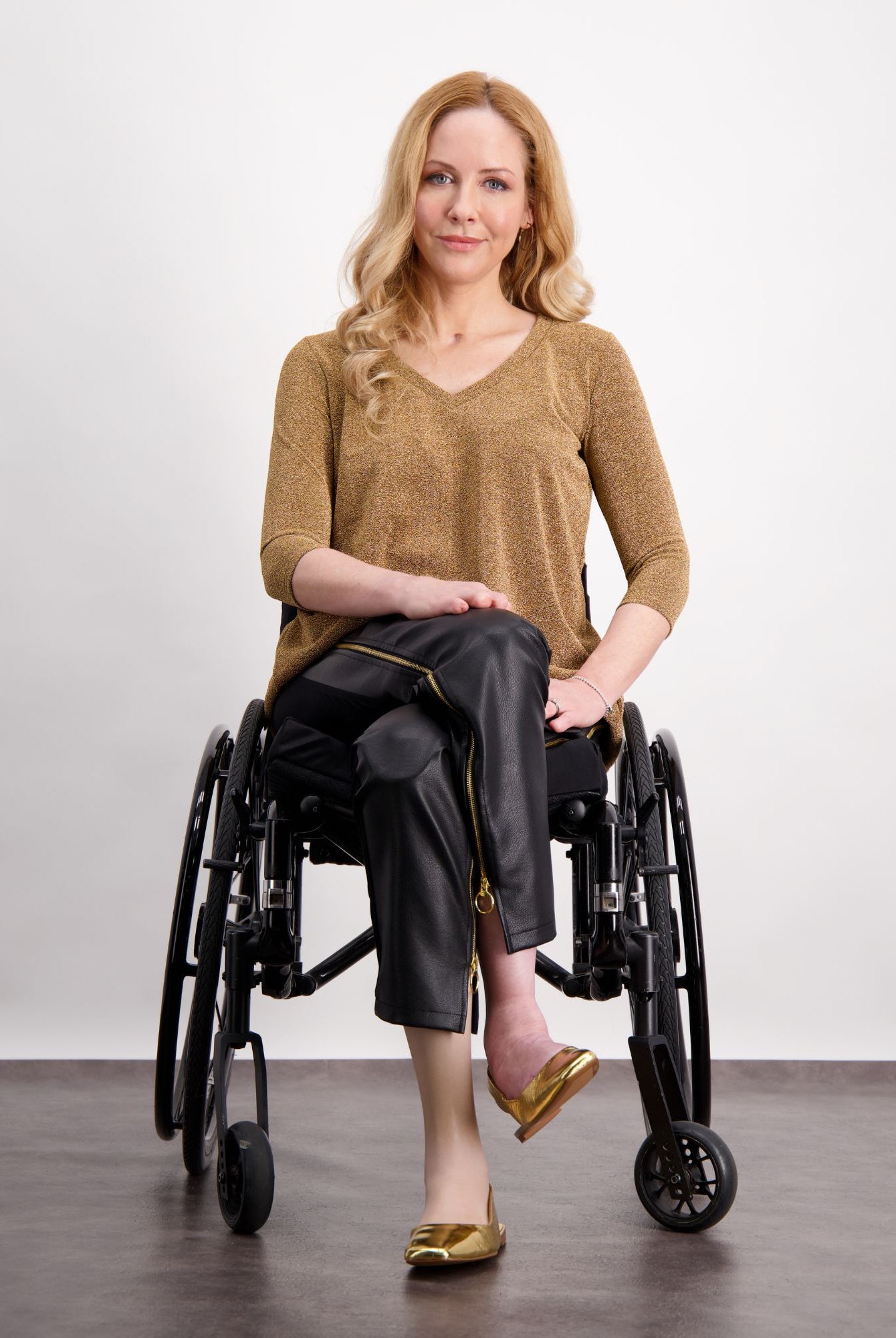 Suzie Q Seated Pants shown on model in wheelchair, featuring black pleather material with gold zippers, paired with metallic gold v-neck top and matching flats. Adaptive design visible with spacious fit and accessible zipper details against white studio background.