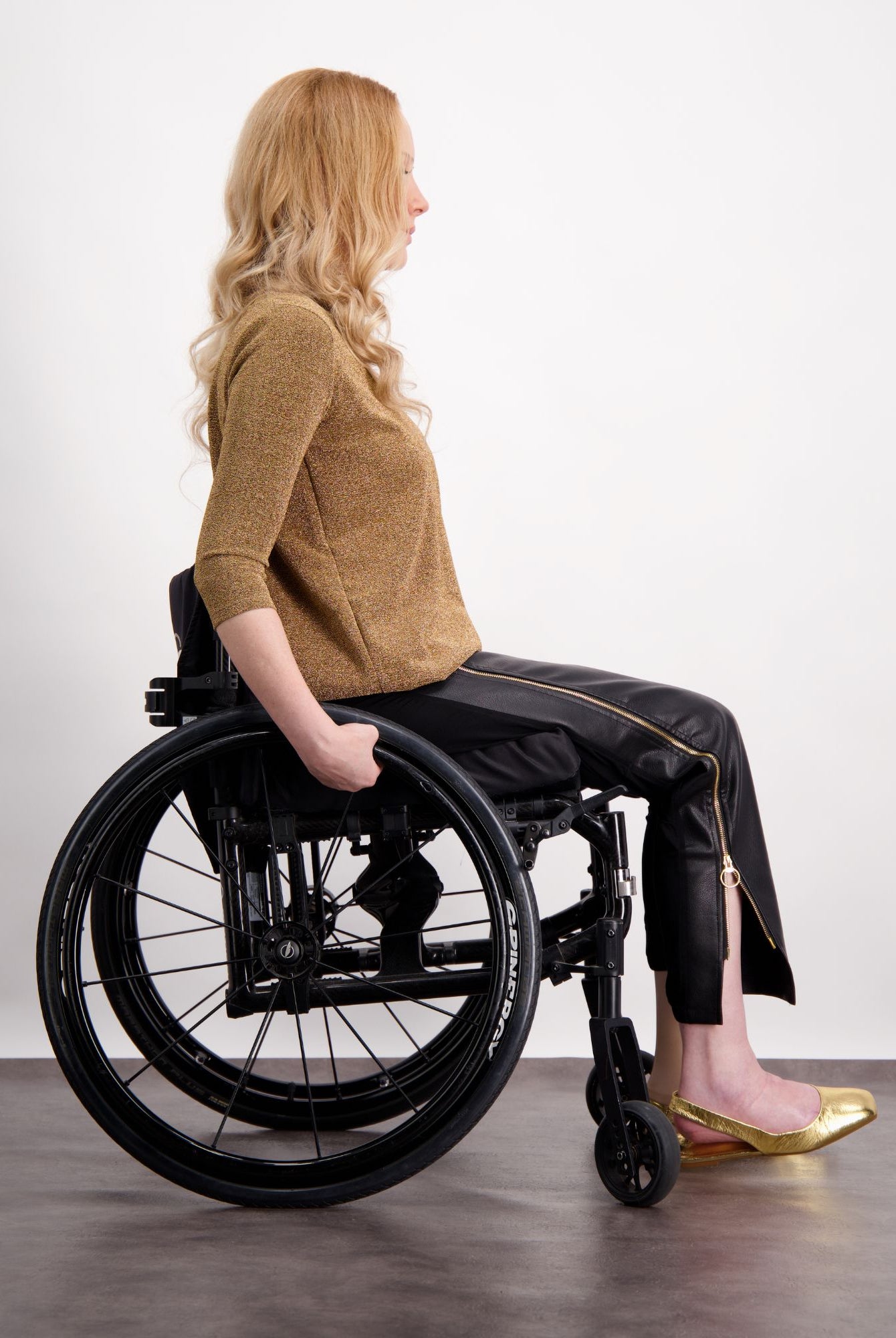 Suzie Q Seated Pants shown on a model in a wheelchair, featuring a black adaptive design with gold accents, paired with a brown sweater and metallic flats. Side view demonstrates seamless fit and wheelchair-friendly construction.