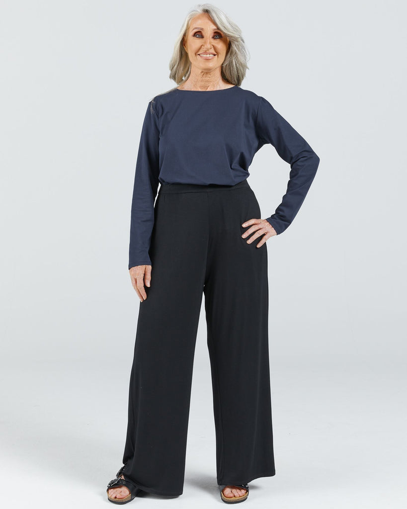 Image is of a female wearing a pair of black bamboo wide leg pants, a navy long sleep leaf-back t-shirt and black leather slippers.  She has grey hair and her hand is resting on her hip. Christina Stephens Australian Adaptive Clothing.