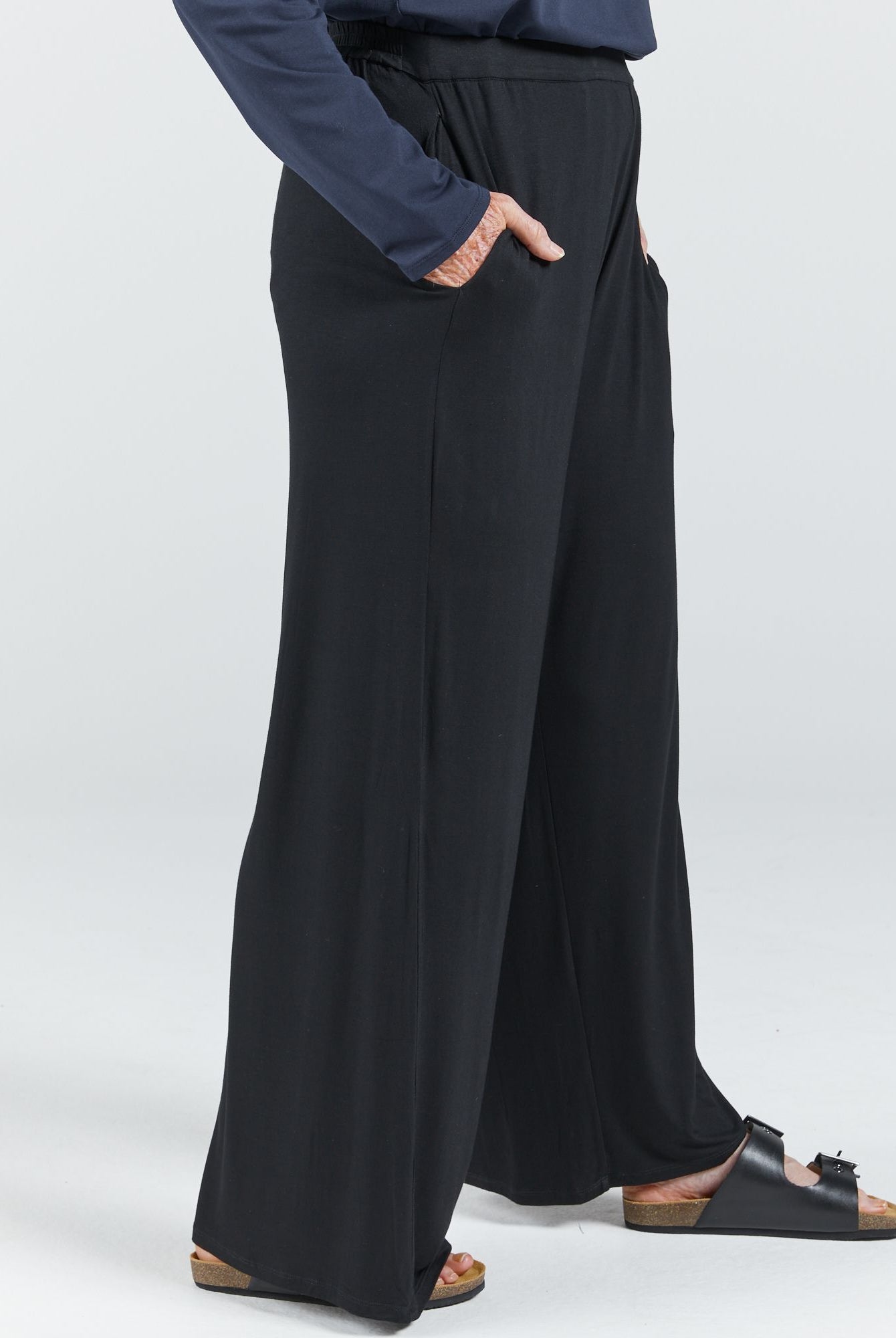Jessie Wide Leg Pant shown from hip to ankle, featuring a flowing black silhouette with side pockets and relaxed fit. Model wears dark sandals. Navy blue top partially visible. Photographed against light grey background, highlighting the pants' elegant drape and comfort-focused design.