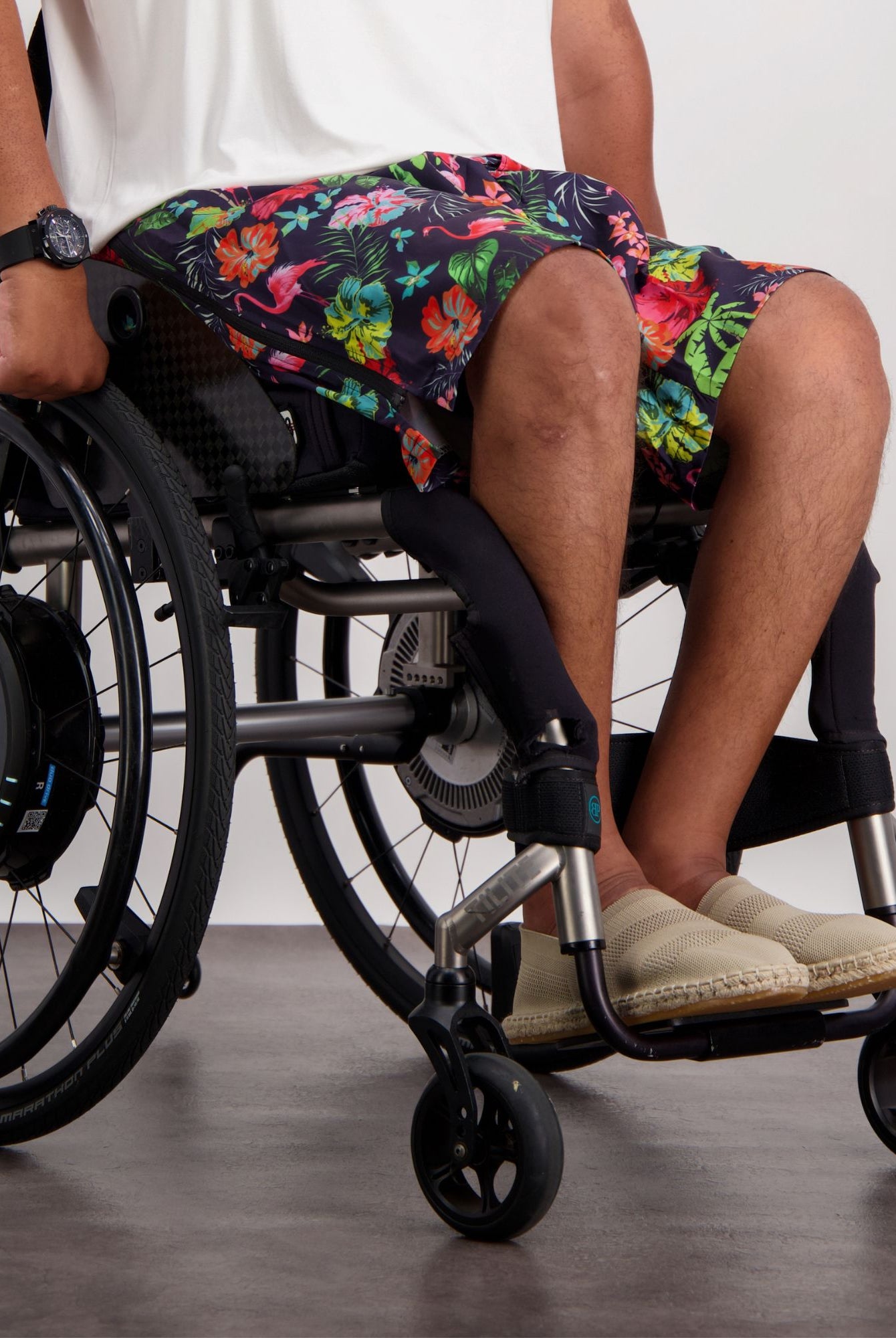 Lindsay Seated Board Shorts shown on a wheelchair user, featuring vibrant tropical floral print in pink, orange, and green on navy background, with comfortable fit designed for seated wear, paired with casual beige footwear and digital watch accessory.
