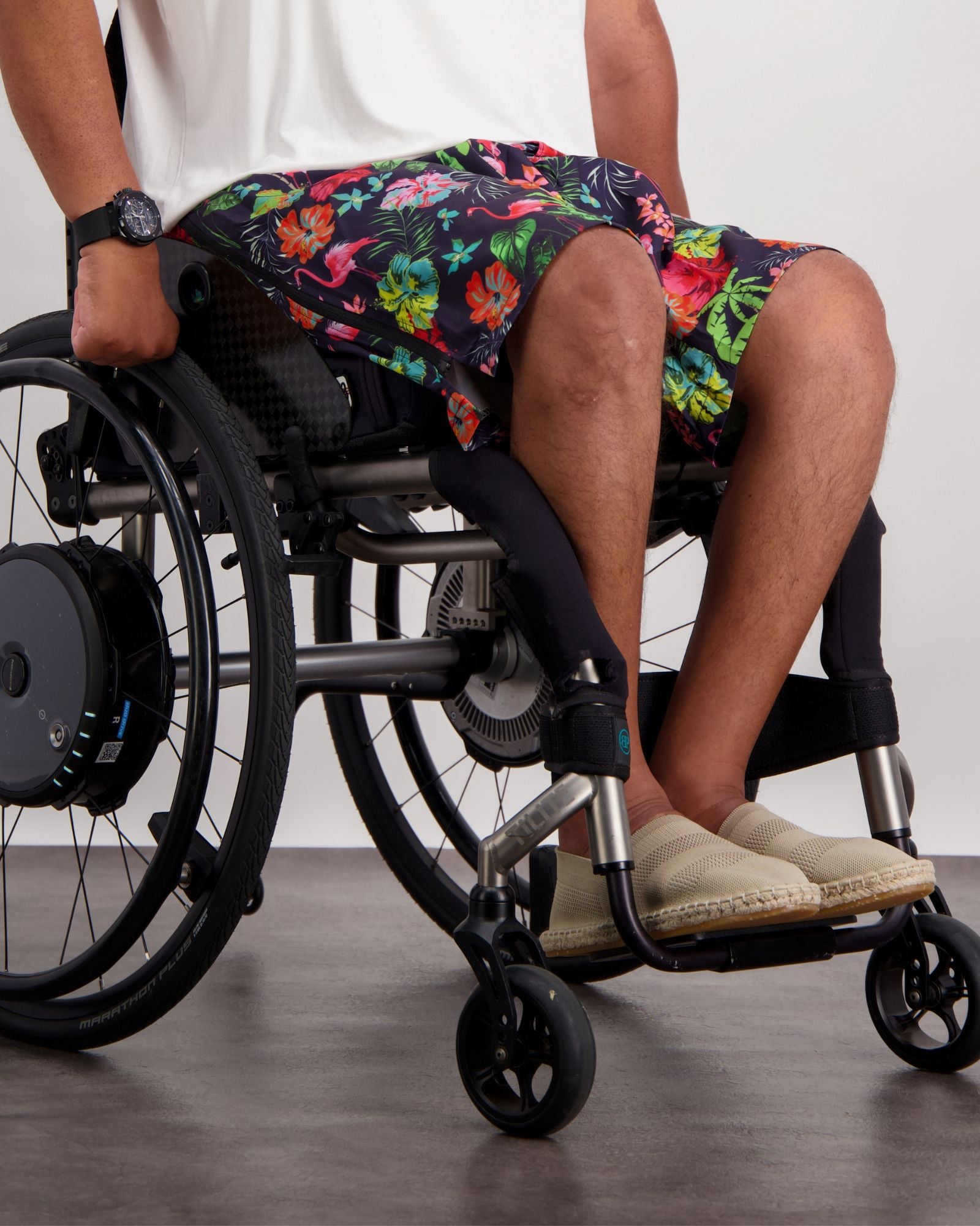 Lindsay Seated Board Shorts shown on a wheelchair user, featuring vibrant tropical floral print in pink, orange, and green on navy background, with comfortable fit designed for seated wear, paired with casual beige footwear and digital watch accessory.