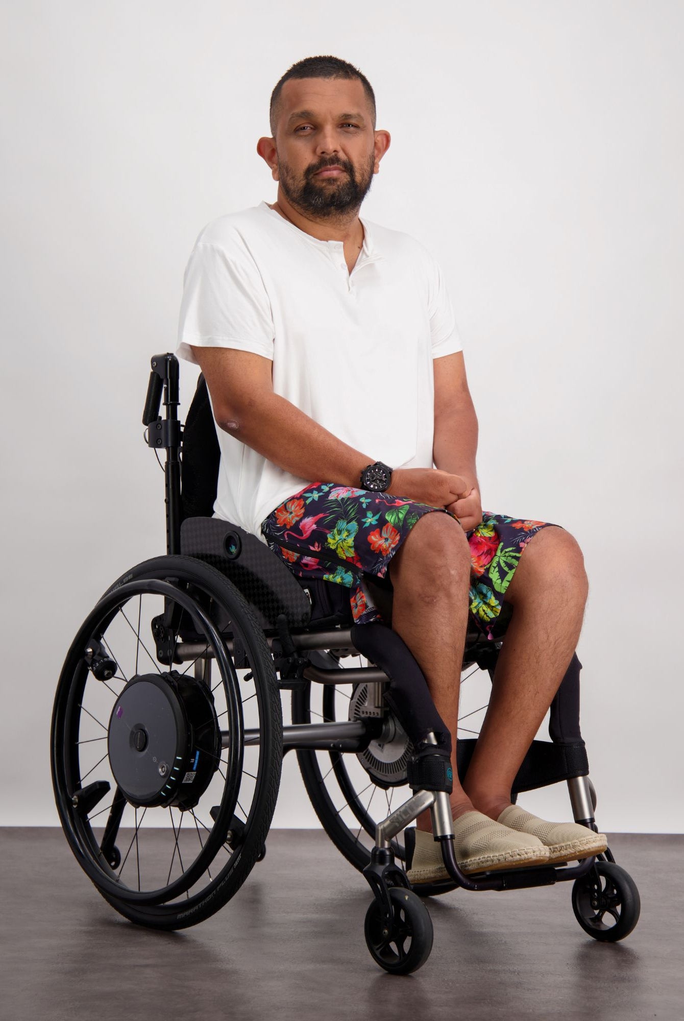 Model wearing Lindsay Seated Board Shorts in vibrant floral print, sitting in a modern wheelchair against a white background, paired with a casual white t-shirt, demonstrating adaptive fashion comfort and style.