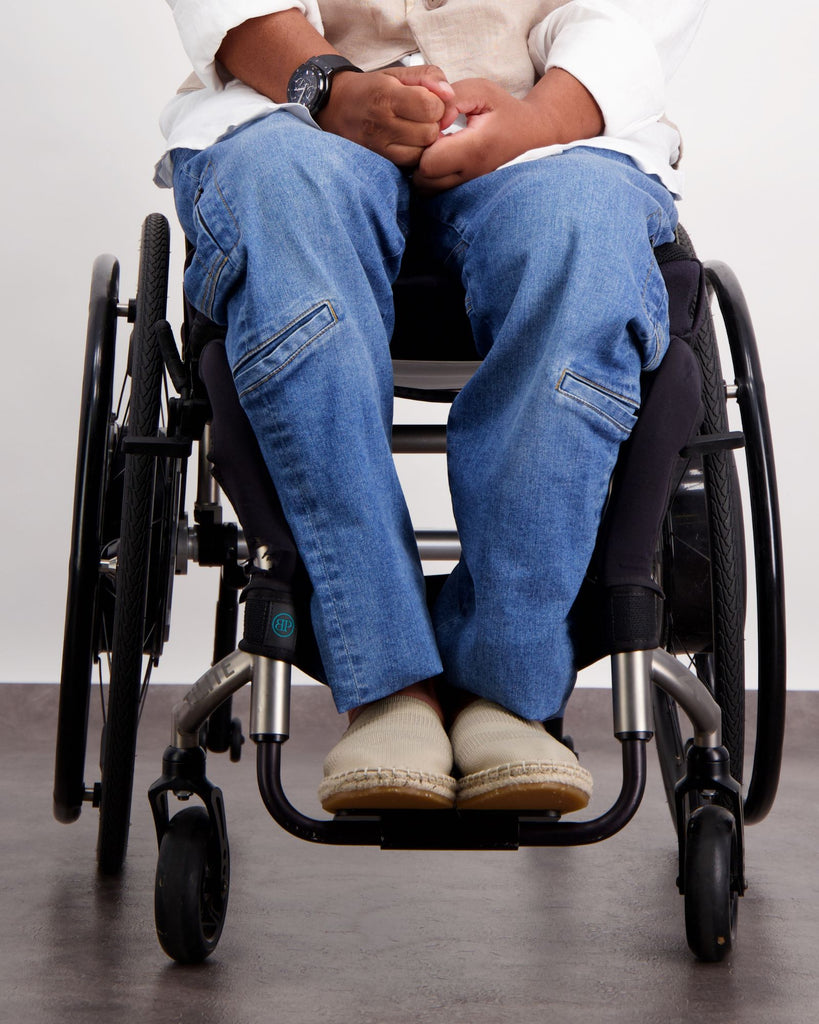 Male with dark skin and dark hair is sitting in a wheelchair wearing a white linen shirt, sand coloured linen vest, blue jeans and sand coloured loafer shoes. Christina Stephens Adaptive Clothing Australia. 