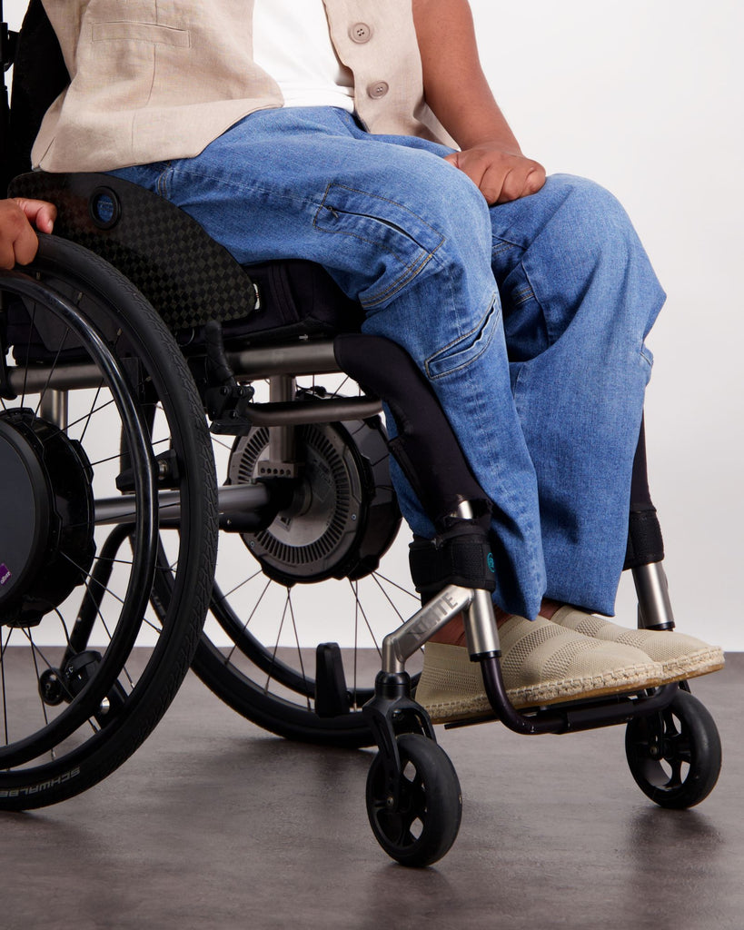 Male with dark skin and dark hair is sitting in a wheelchair wearing a white linen shirt, sand coloured linen vest, blue jeans and sand coloured loafer shoes. Christina Stephens Adaptive Clothing Australia. 