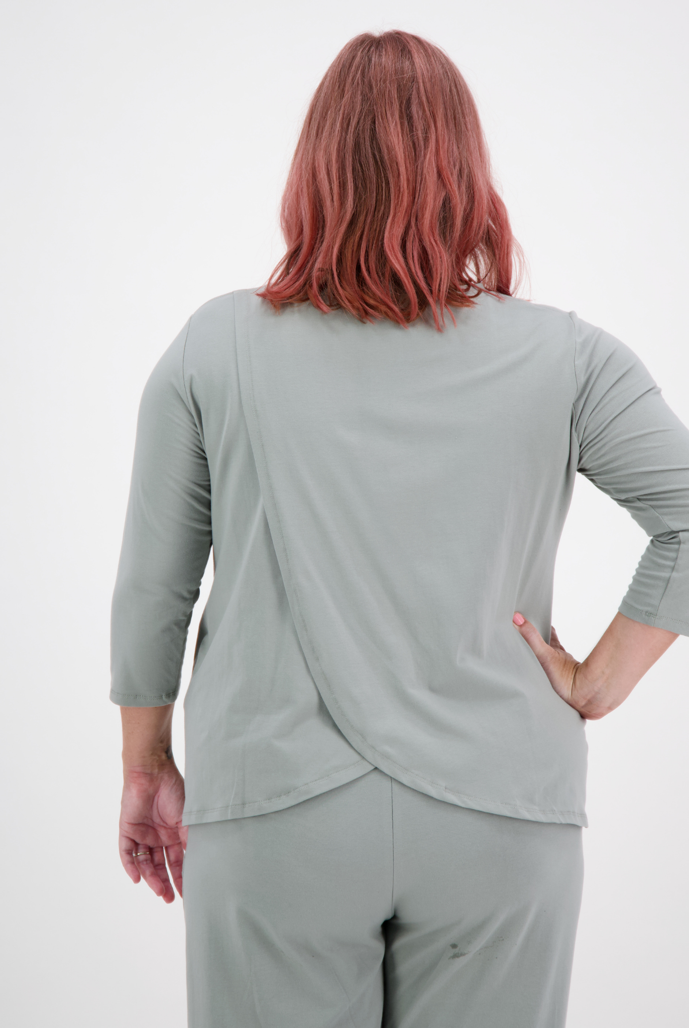 Back view of 3/4 Sleeve Leaf Back T-Shirt in soft sage green, showcasing distinctive overlapping leaf design at lower back, styled with matching pants. Rose-colored hair model demonstrates adaptive stretch and elegant drape of the garment.