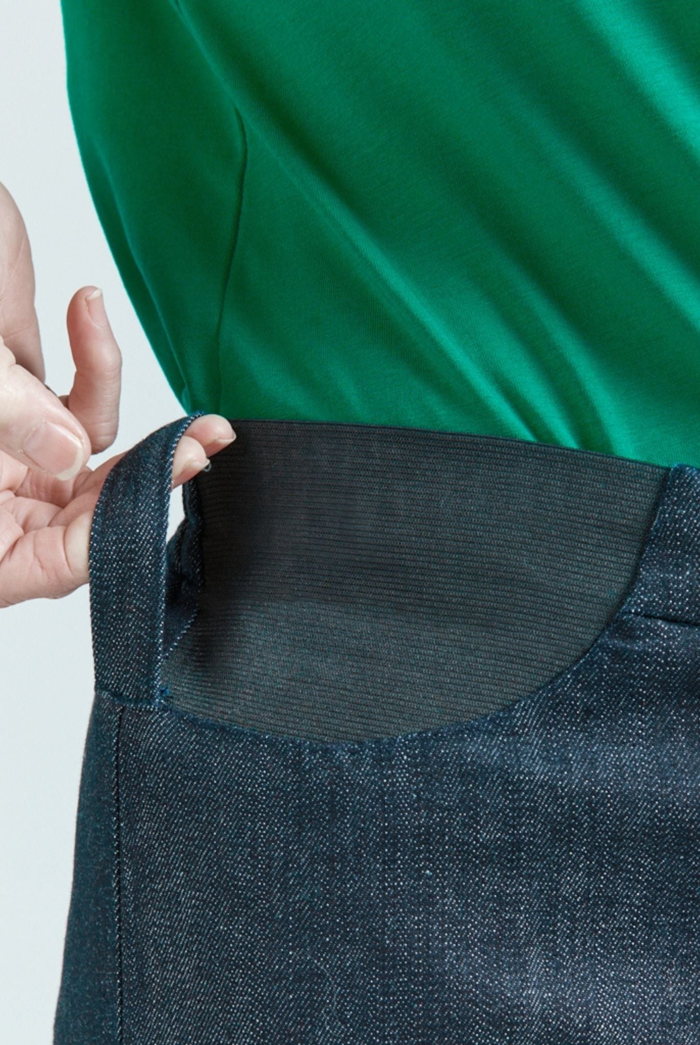 Seated Stretch Pant featuring an elastic waistband detail in dark denim, shown against an emerald green top. Close-up view highlighting the adaptable stretch panel design that prevents riding up while seated, demonstrating the pants' functional yet stylish construction.