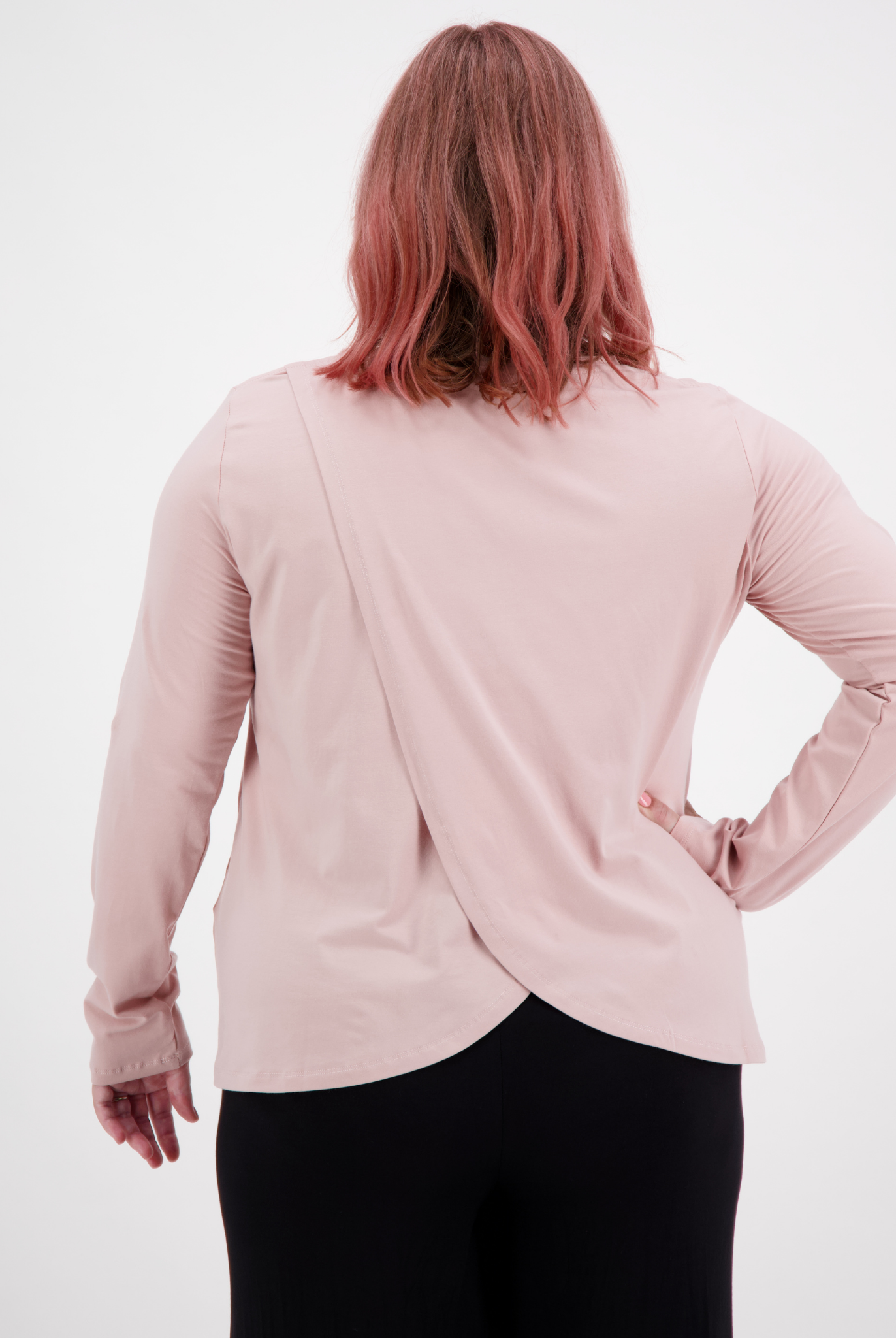 Long Sleeve Leaf Back T-Shirt in soft pink featuring unique cross-over back design, shown from rear view against white background. Stylish casual top paired with black bottoms, demonstrating the elegant drape and overlap detail of the back panel.