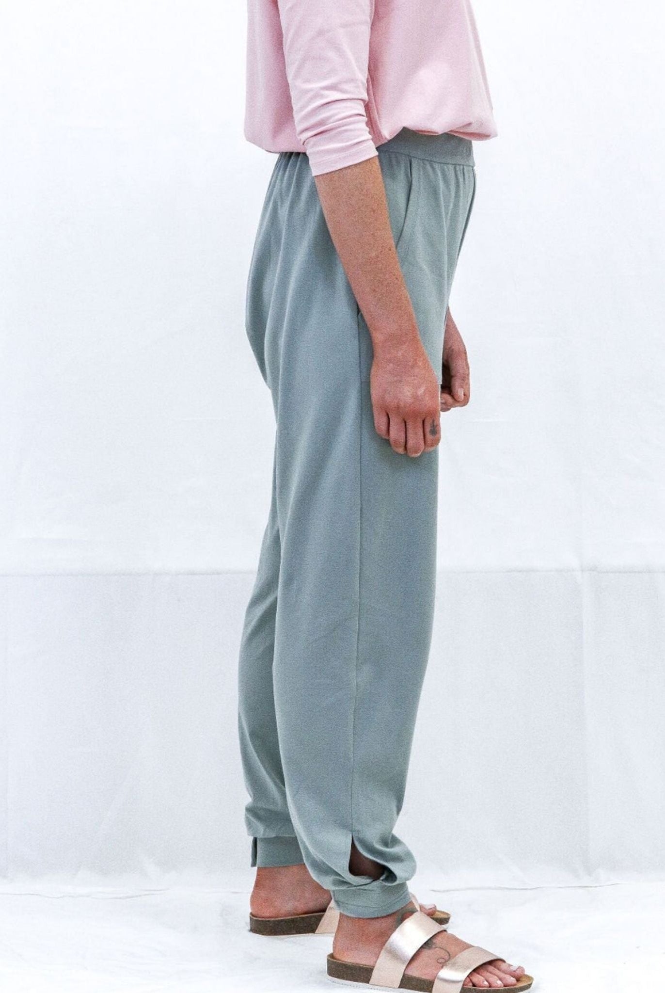 Tapered Track Pants with Adjustable Cuff shown in sage green color, styled with a pink top. Side view displaying the relaxed fit, elastic waistband, and tapered leg design with adjustable cuffs at ankles, paired with casual sandals.
