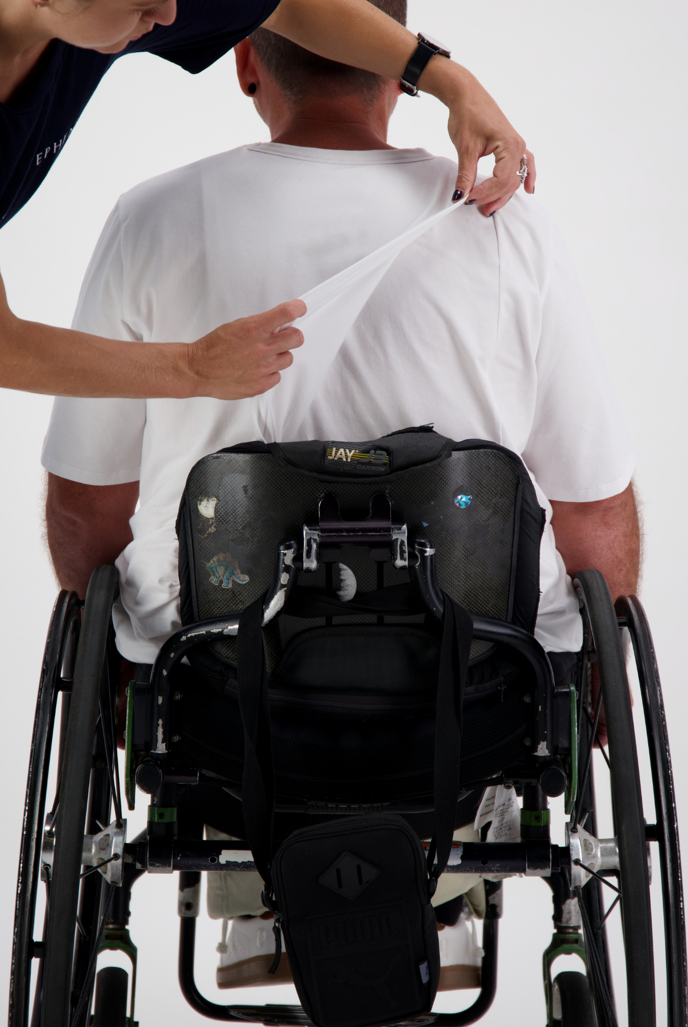 Short Sleeve Prism Back T-Shirt being demonstrated on a wheelchair user, showing easy removal feature with caregiver assistance. White shirt against white background with black wheelchair and adaptive backpack visible.