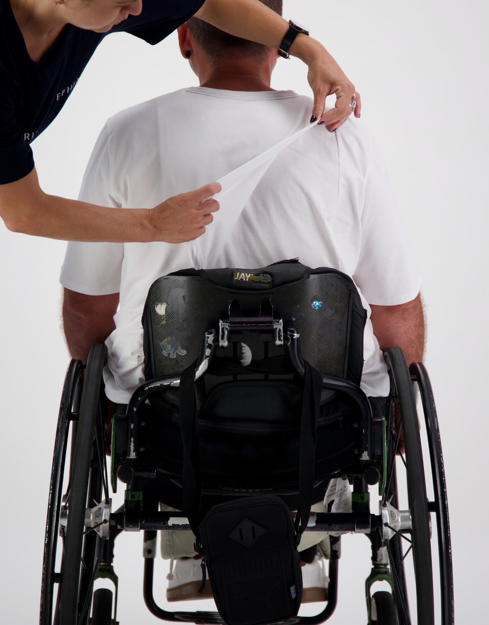 Short Sleeve Prism Back T-Shirt being demonstrated on a wheelchair user, showing easy removal feature with caregiver assistance. White shirt against white background with black wheelchair and adaptive backpack visible.