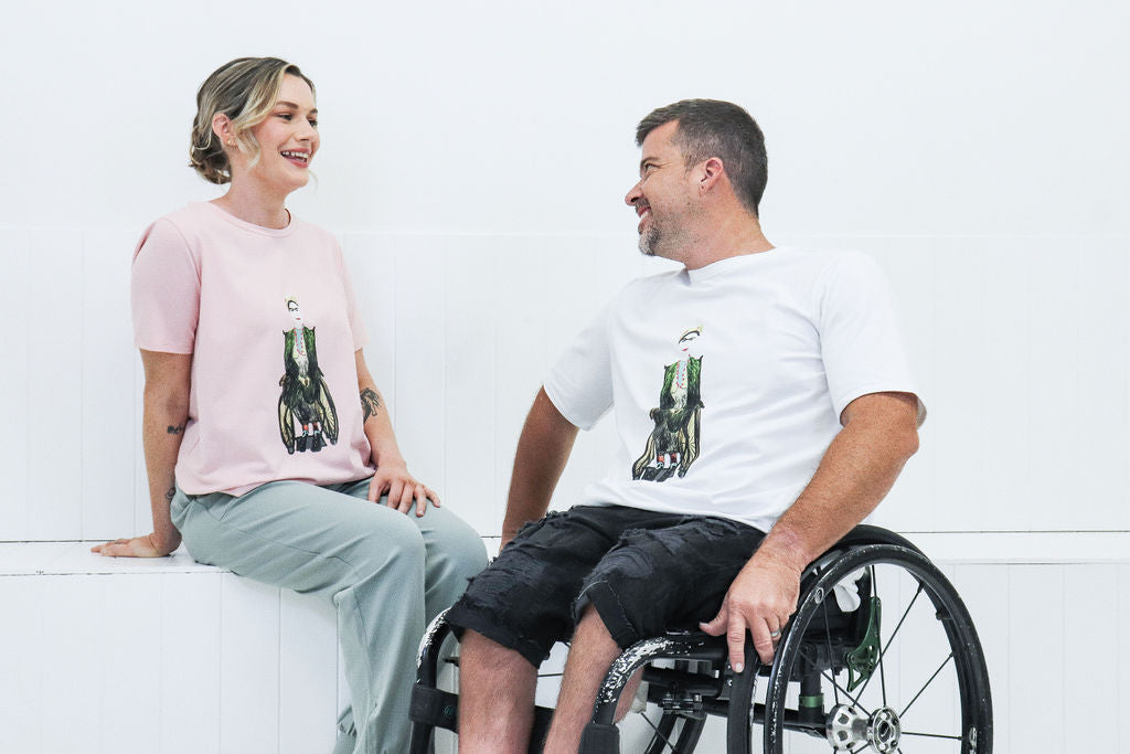 A blonde fair-skinned woman wearing a pink shirt with "Frida" artwork printed on the front, sits on a white bench smiling at a fair-skinned man sitting in a wheelchair next to her. He is wearing a white t-shirt with the same artwork and black shorts. 