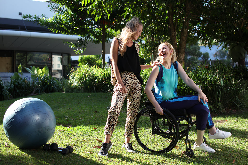Two girls outside on the grass smiling at each other. On the left we have an exercise ball. The first girl is wearing a black top and leopard pants standing. The second girl is wearing a blue watercolour top and navy watercolour pants in a wheelchair.