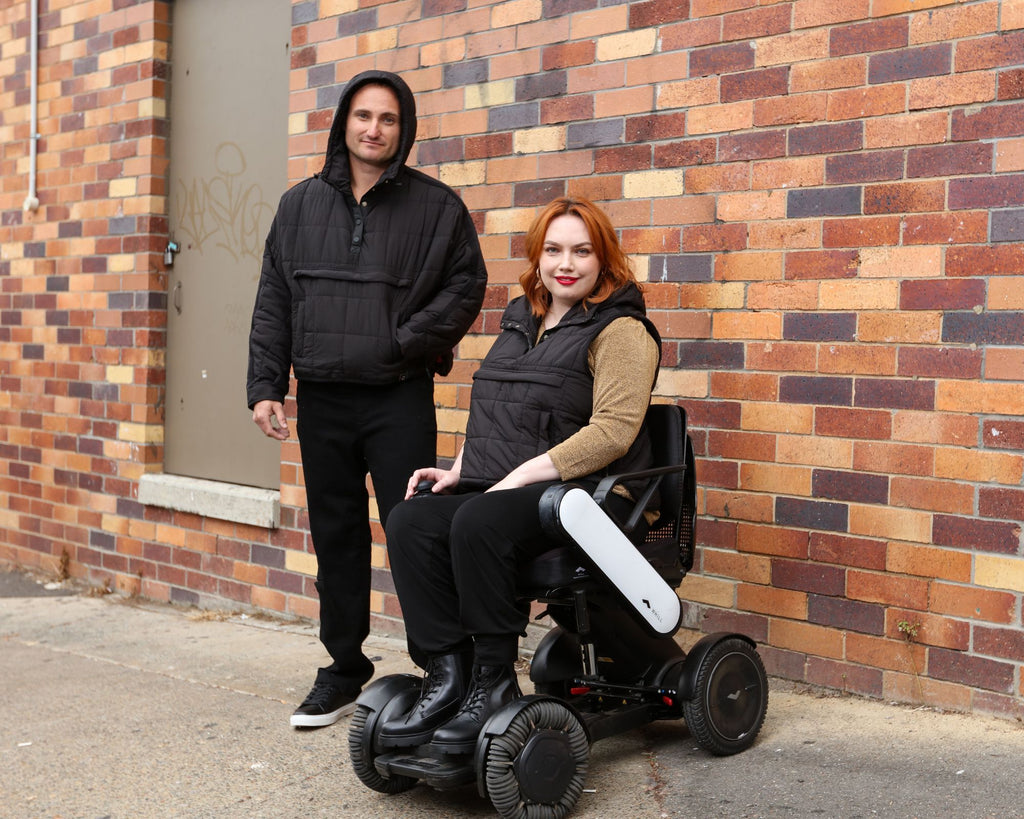 Standing male and seated female wearing puffer jackets and vests in front of brick wall. Christina Stephens Adaptive Clothing Australia. 
