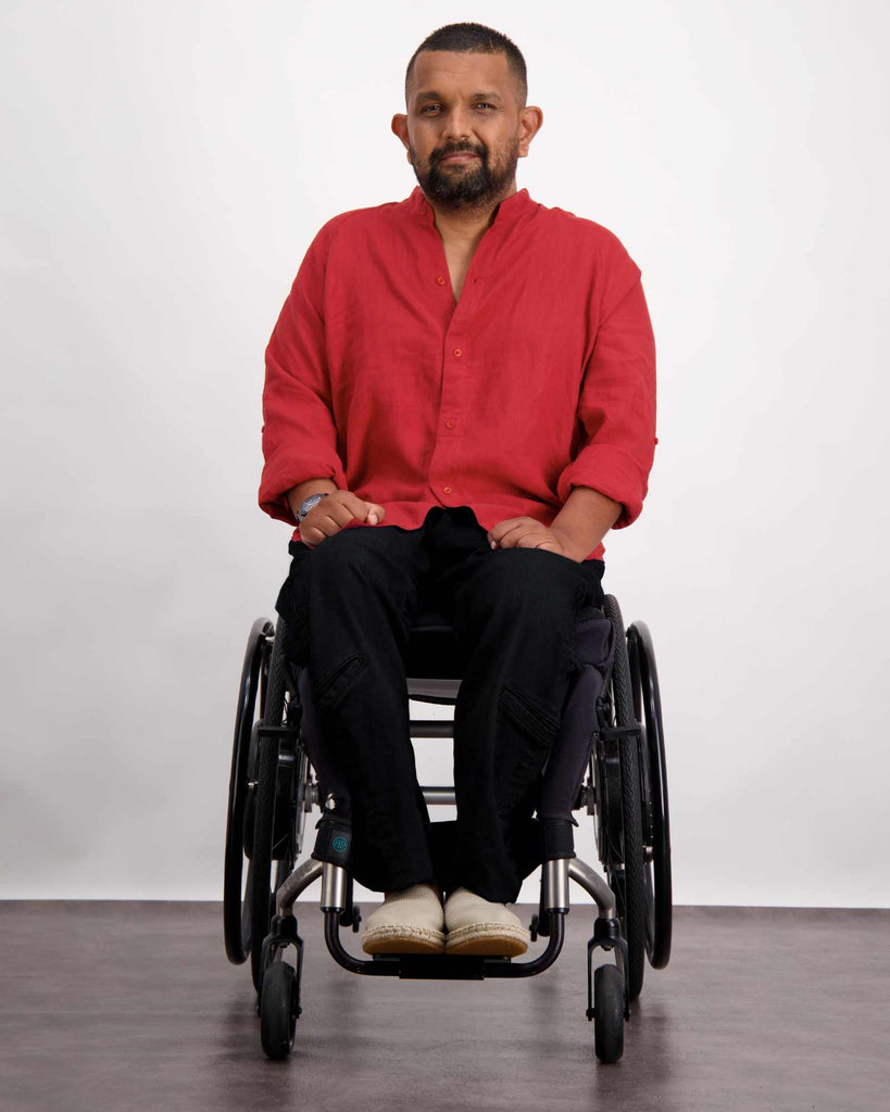 Male with dark skin and dark hair is sitting in a wheelchair wearing a RED linen shirt, black jeans and sand coloured loafer shoes. Christina Stephens Adaptive Clothing Australia.