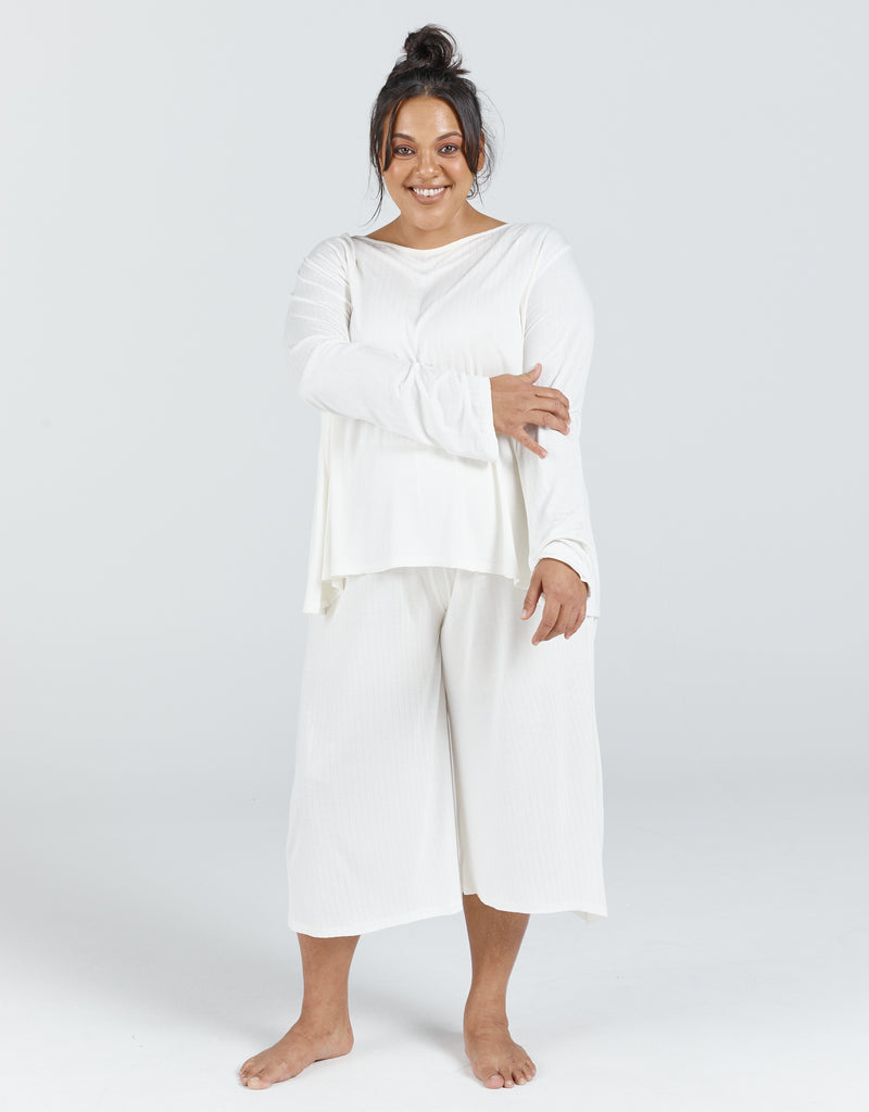 Standing female with brunette hair up in a bun is wearing an ivory bamboo leaf back top with long sleeves. She is also wearing a cropped wide leg pant made from the same fabric. Christina Stephens Adaptive Clothing Australia