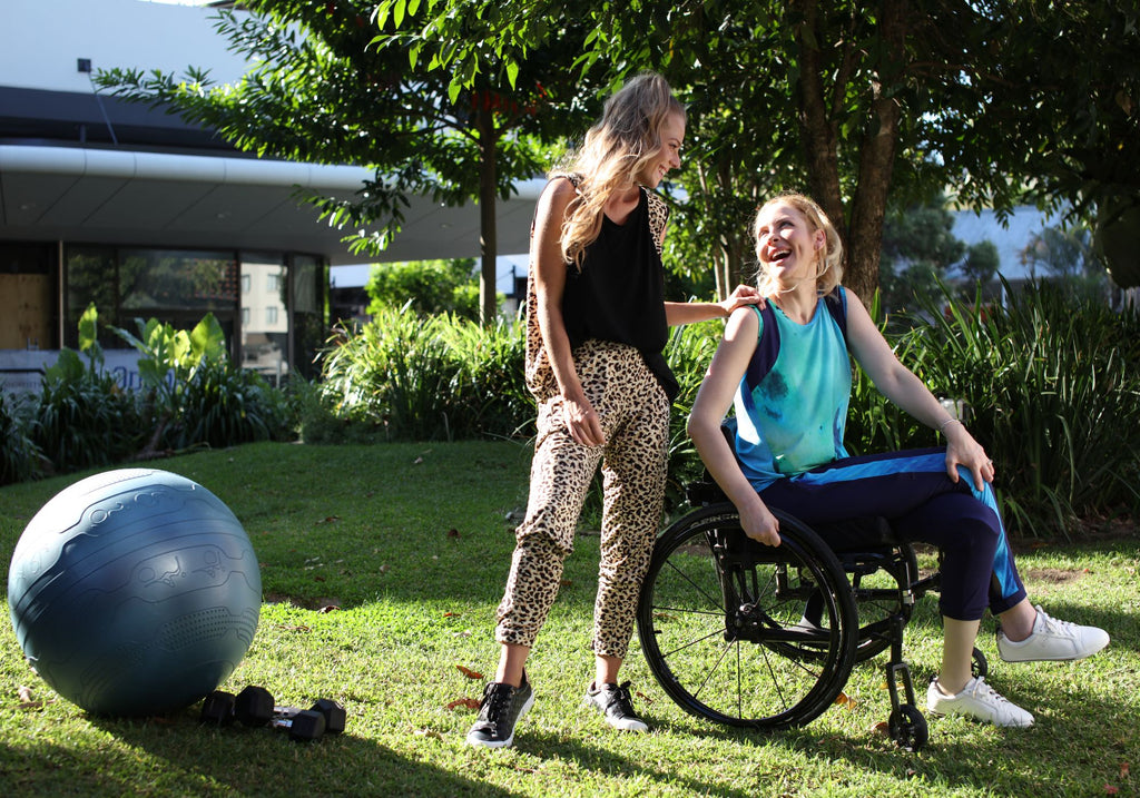 A blonde woman standing wearing leopard print pants and a black singlet laughs with a blonde woman in a wheelchair wearing ocean blue singlet and blue pants.