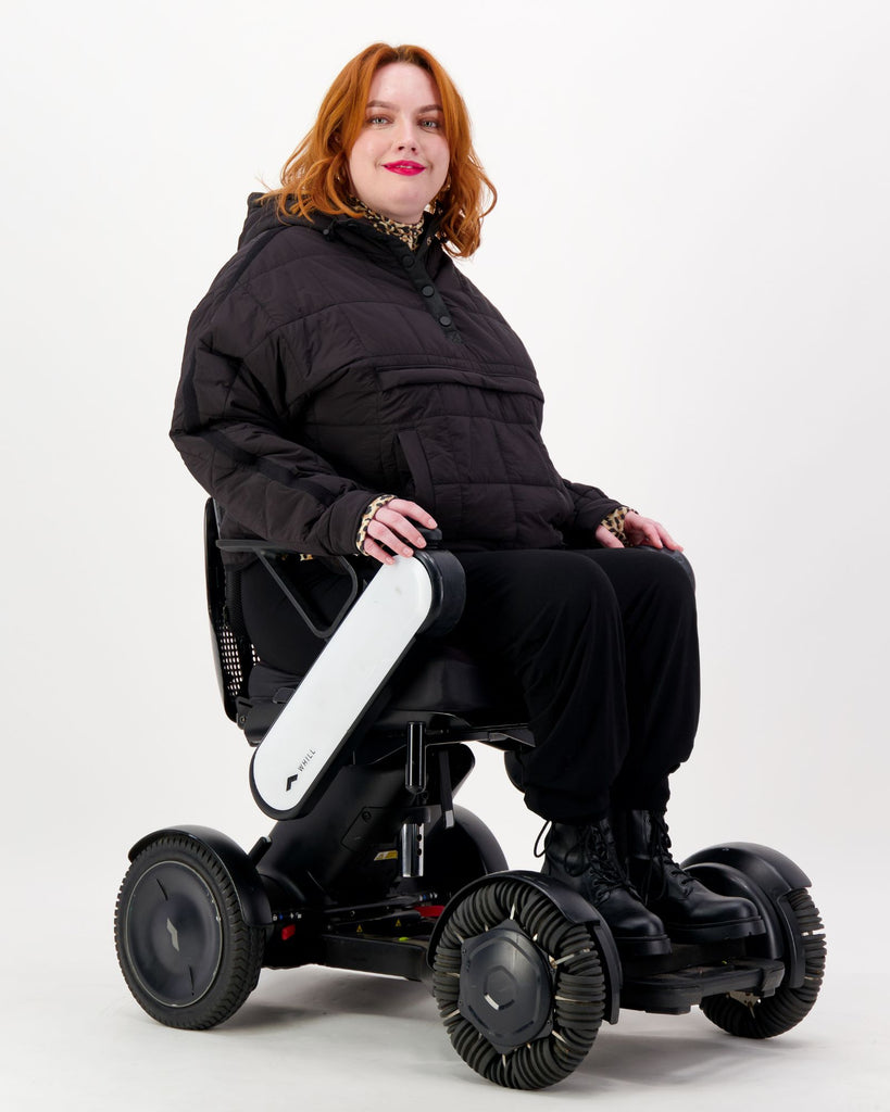 Seated female in a power chair is wearing a black puffer jacket over black pants. Christina Stephens Adaptive Clothing Australia.