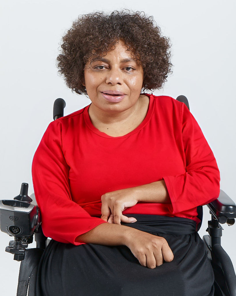 Woman with dark hair and skin is sitting in a wheelchair wearing a blue red bright coloured 3/4 sleeve t-shirt. Christina Stephens Adaptive Clothing Australia.