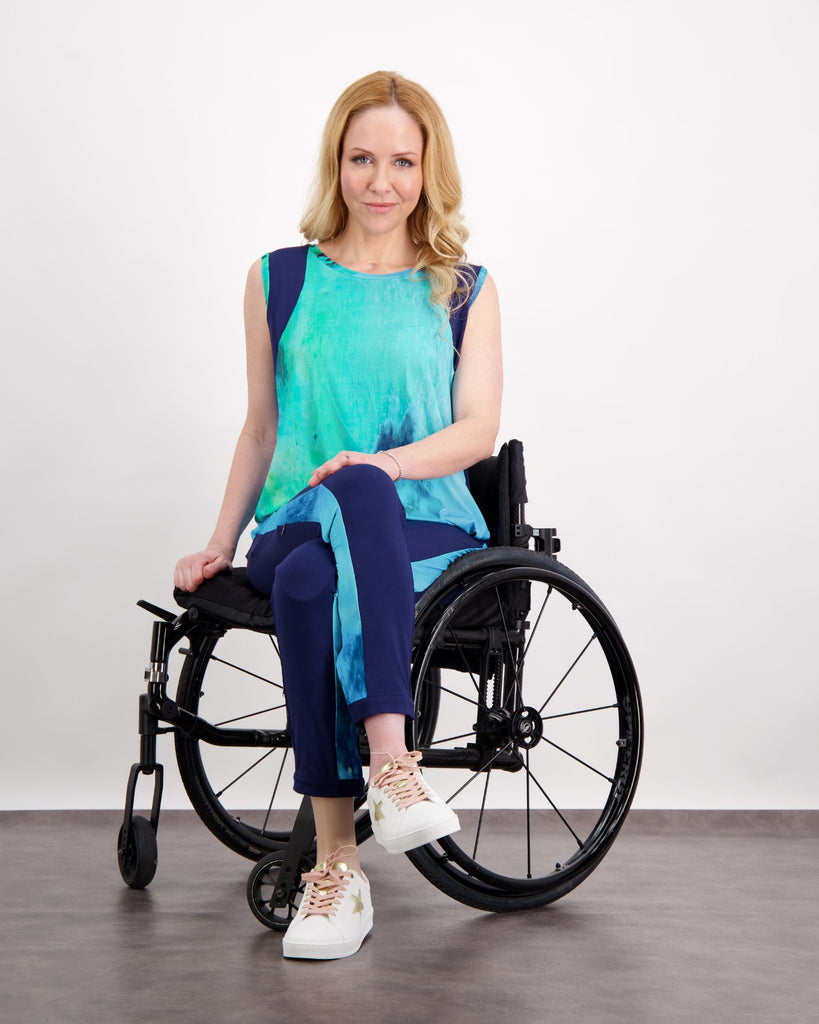 Blonde female sitting in a manual wheelchair wears an aqua blue print top and matching blue and navy physio pants with white sneakers. Christina Stephens Adaptive Clothing Australia. 