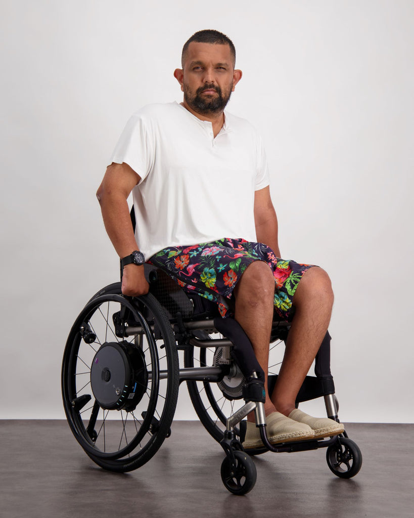 A male in a wheelchair with dark skin tone and dark hair is wearing a white bamboo a-line t-shirt with three small buttons at the neck and brightly coloured board shorts. Christina Stephens Australian Adaptive Clothing.