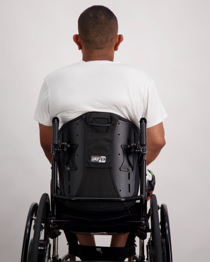 A male in a wheelchair with dark skin tone and dark hair is wearing a white bamboo a-line t-shirt with three small buttons at the neck and brightly coloured board shorts. Christina Stephens Australian Adaptive Clothing.