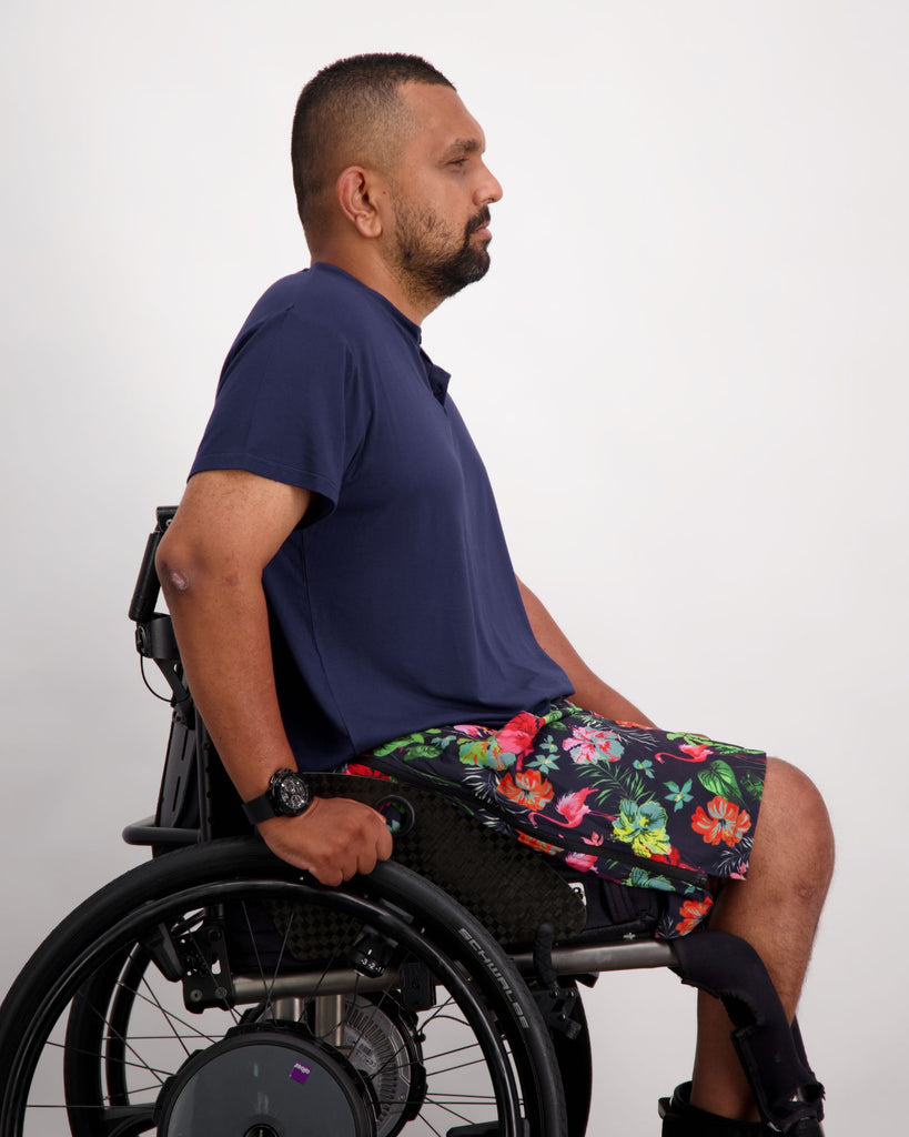 A male in a wheelchair with dark skin tone and dark hair is wearing a navy bamboo a-line t-shirt with three small buttons at the neck and brightly coloured board shorts. Christina Stephens Australian Adaptive Clothing.