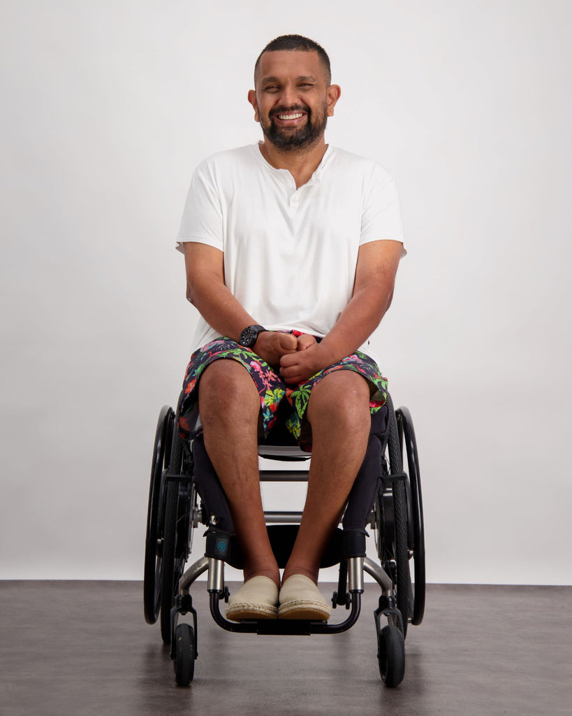 A male in a wheelchair with dark skin tone and dark hair is wearing a white bamboo a-line t-shirt with three small buttons at the neck and brightly coloured board shorts. Christina Stephens Australian Adaptive Clothing.