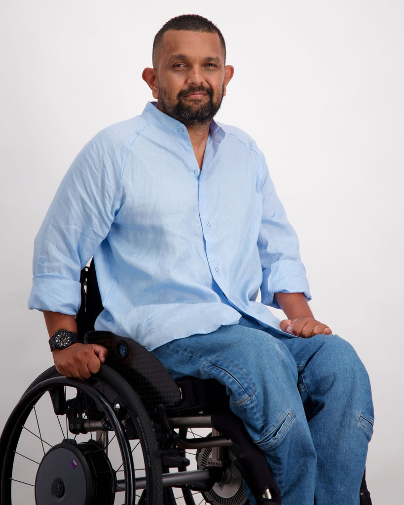 A male in a wheelchair with dark skin tone and dark hair is wearing a light blue linen shirt and a pair of blue denim adaptive jeans. Christina Stephens Australian Adaptive Clothing.