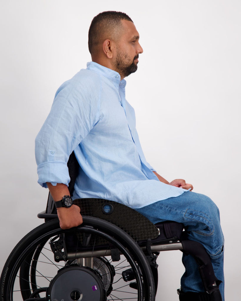 A male in a wheelchair with dark skin tone and dark hair is wearing a light blue linen shirt and a pair of blue denim adaptive jeans. Christina Stephens Australian Adaptive Clothing.