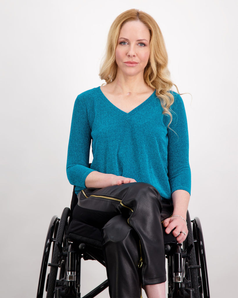 Blonde female sitting in a manual wheelchair is wearing a blue 3/4 sleeve, V-neck glitter top and black vegan leather pants. Christina Stephens Adaptive Clothing Australia.