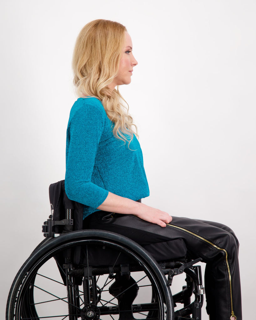 Blonde female sitting in a manual wheelchair is wearing a blue 3/4 sleeve, V-neck glitter top and black vegan leather pants. Christina Stephens Adaptive Clothing Australia.