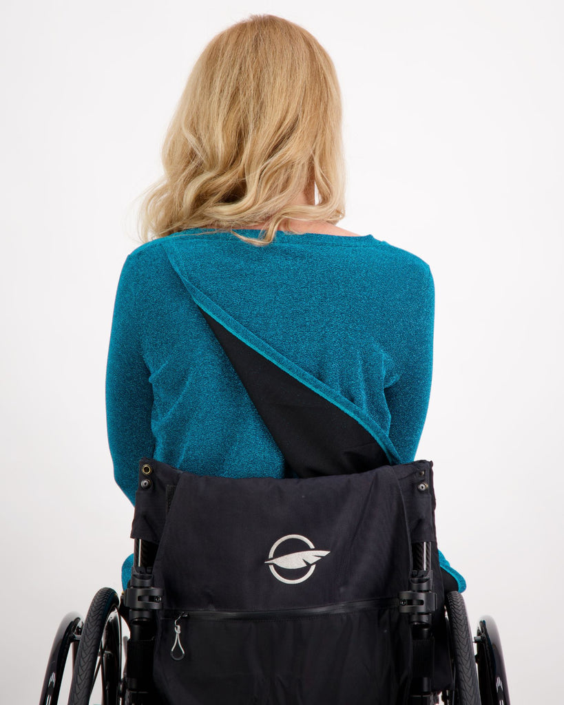 Blonde female sitting in a manual wheelchair is wearing a blue 3/4 sleeve, V-neck glitter top and black vegan leather pants. She is facing the back showing the leaf back design of the top. Christina Stephens Adaptive Clothing Australia.