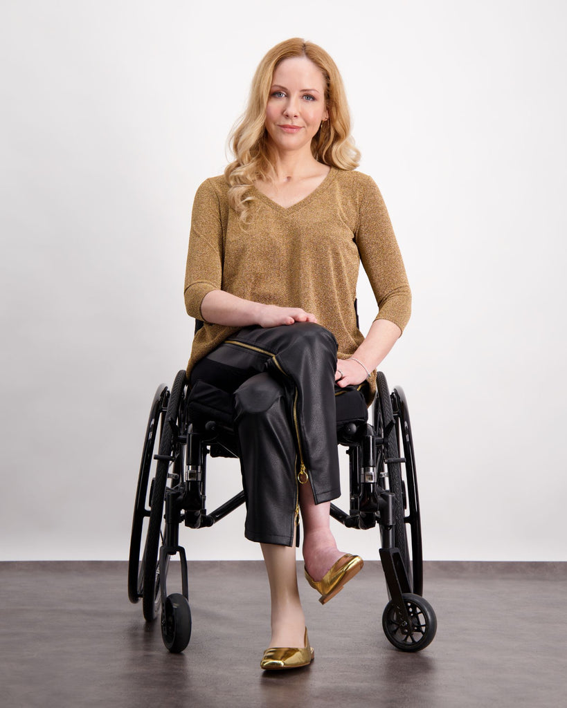 A blonde female is sitting in a wheelchair wearing a gold glitter top with 3/4 length sleeve, black vegan leather pants with gold zips and gold slip on shoes. Christina Stephens Adaptive Clothing Australia. 