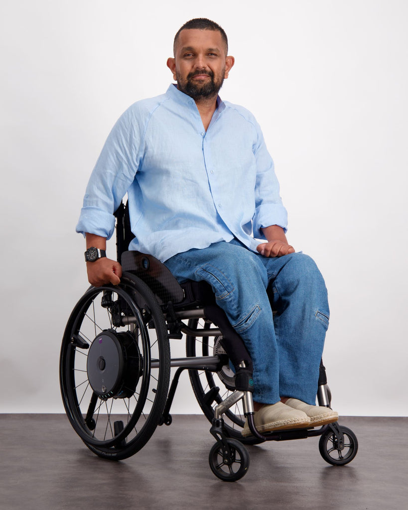 Male sitting in a wheelchair with dark skin and short dark hair also has a short beard. He is wearing a light blue linen shirt, blue adaptive denim jeans and sand coloured shoes. Christina Stephens Adaptive Clothing Australia.