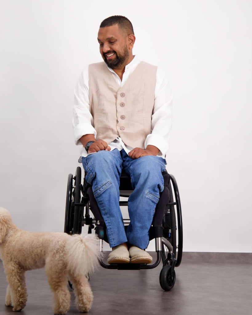 A male with dark skin and dark hair is sitting in a wheelchair wearing a beige vest, white shirt and blue jeans. There is a white poodle dog in the frame. Christina Stephens Adaptive Clothing Australia.