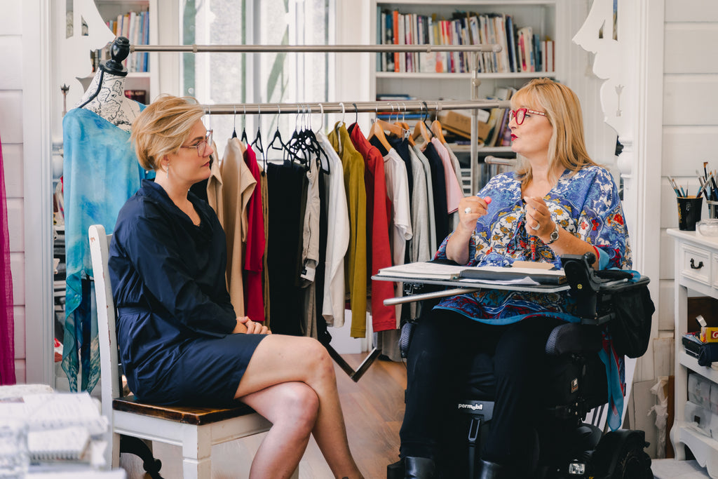 Image of two women (Jessie and Carol) sitting in front of a rack of clothing talking to each other. Christina Stephens Adaptive Clothing Australia.