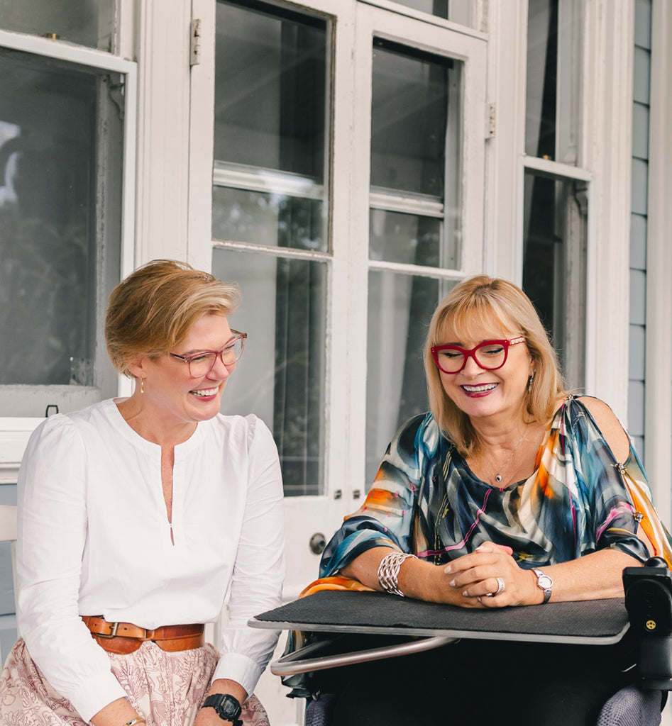 Jessie and Carol sitting on a front Verandah laughing at a joke. Jessie has light coloured short hair and is wearing pink glasses and a white shirt. Carol has shoulder length blonde hair and is wearing red glasses and a green silk kaftan. Christina Stephens Adaptive Clothing Australia.