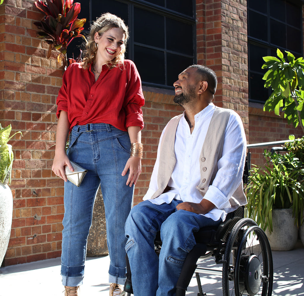 Bella standing, laughing wearing a red linen shirt and blue jeans, Dinesh sitting in a wheelchair smiling up to Bella wearing a white linen shirt, bone waistcoat undone, and blue jeans.
