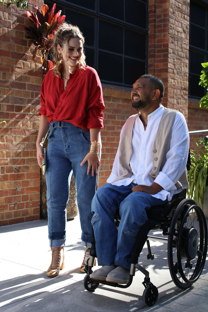 A female with a red shirt and blue jeans stands next to a dark skinned man in a wheelchair wearing a white shirt, beige vest and denim blue jeans. Christina Stephens Adaptive Clothing Australia. 
