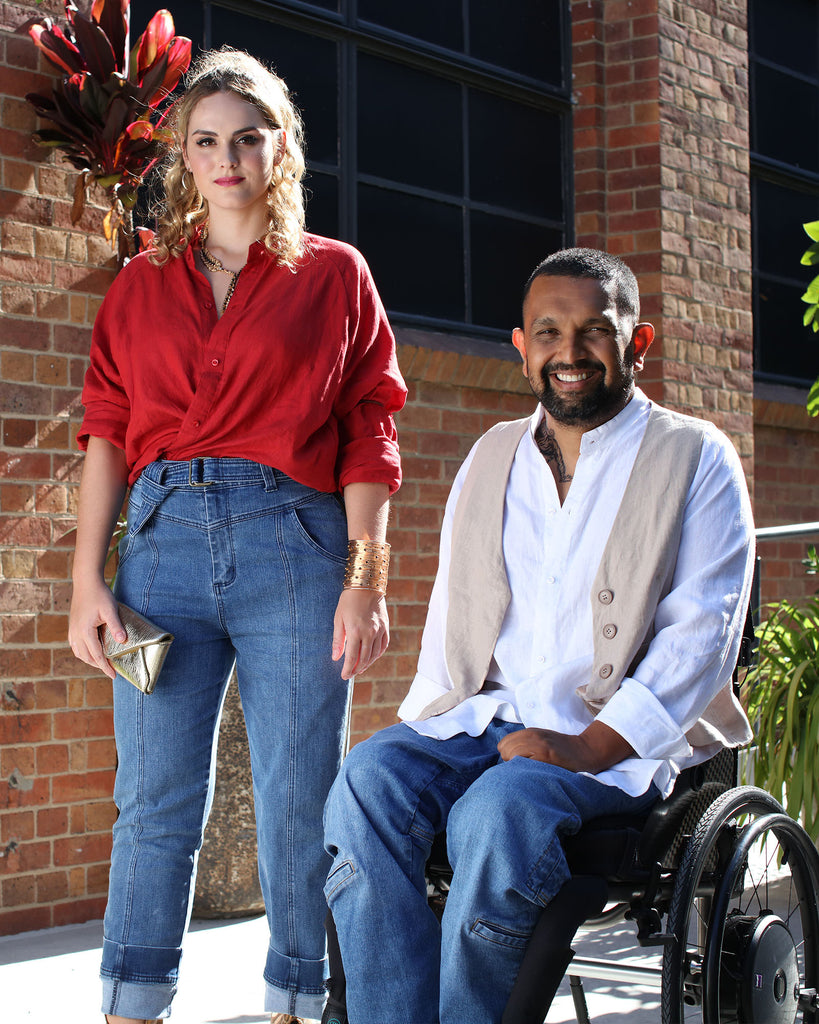 A female wearing a red shirt and blue jeans stands next to a dark skinned man in a wheelchair wearing a white shirt, beige vest and denim blue jeans. Christina Stephens Adaptive Clothing Australia. 