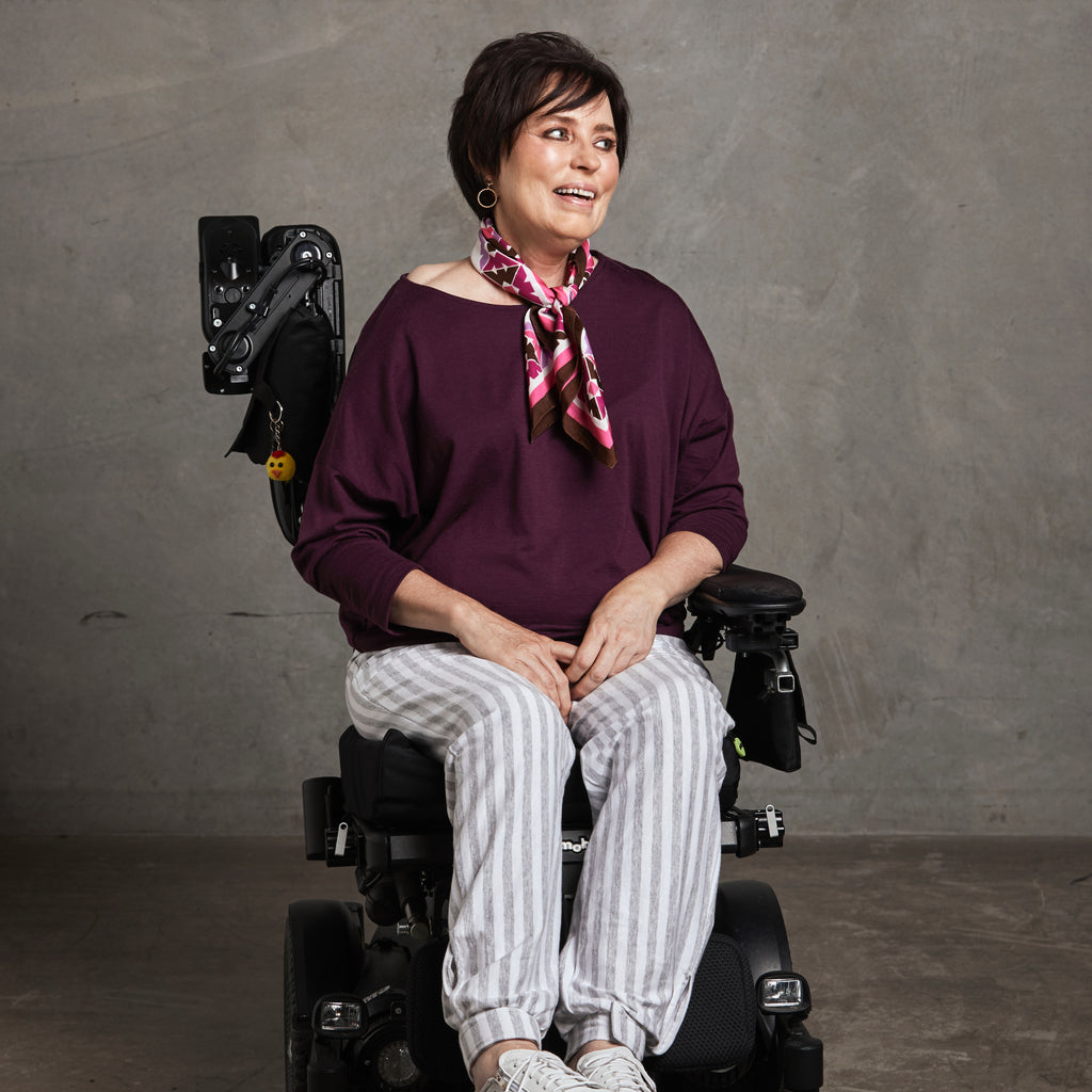 Female in a wheelchair is wearing a purple drop shoulder top and grey and white striped pants. Christina Stephens Adaptive Clothing Australia. 