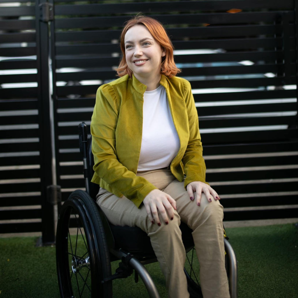 A female wearing a citrus green velvet jacket, white t-shirt and gold linen pants, sits in a wheelchair. She has red hair. Christina Stephens Adaptive Clothing Australia. 