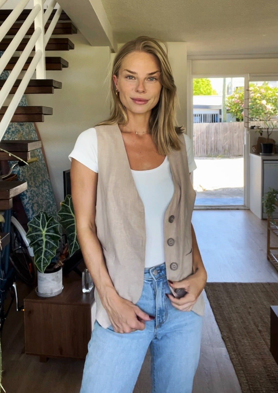 Dean Gender Neutral Waistcoat styled with white t-shirt and light blue jeans, shown in a modern home setting with floating wooden stairs and indoor plants. The beige linen waistcoat features button closures and a classic V-neck cut.