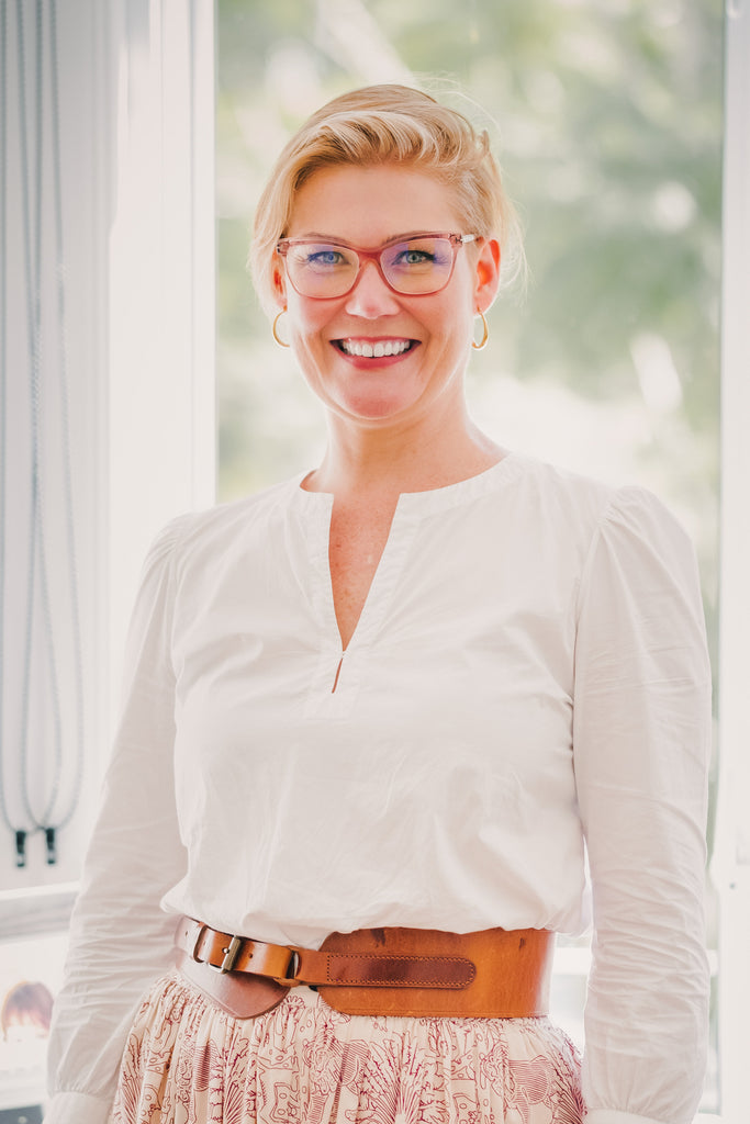 Image of a female (Jessie) standing in front of a window wearing a white shirt and pink glasses. She is smiling. Christina Stephens Adaptive Clothing Australia.